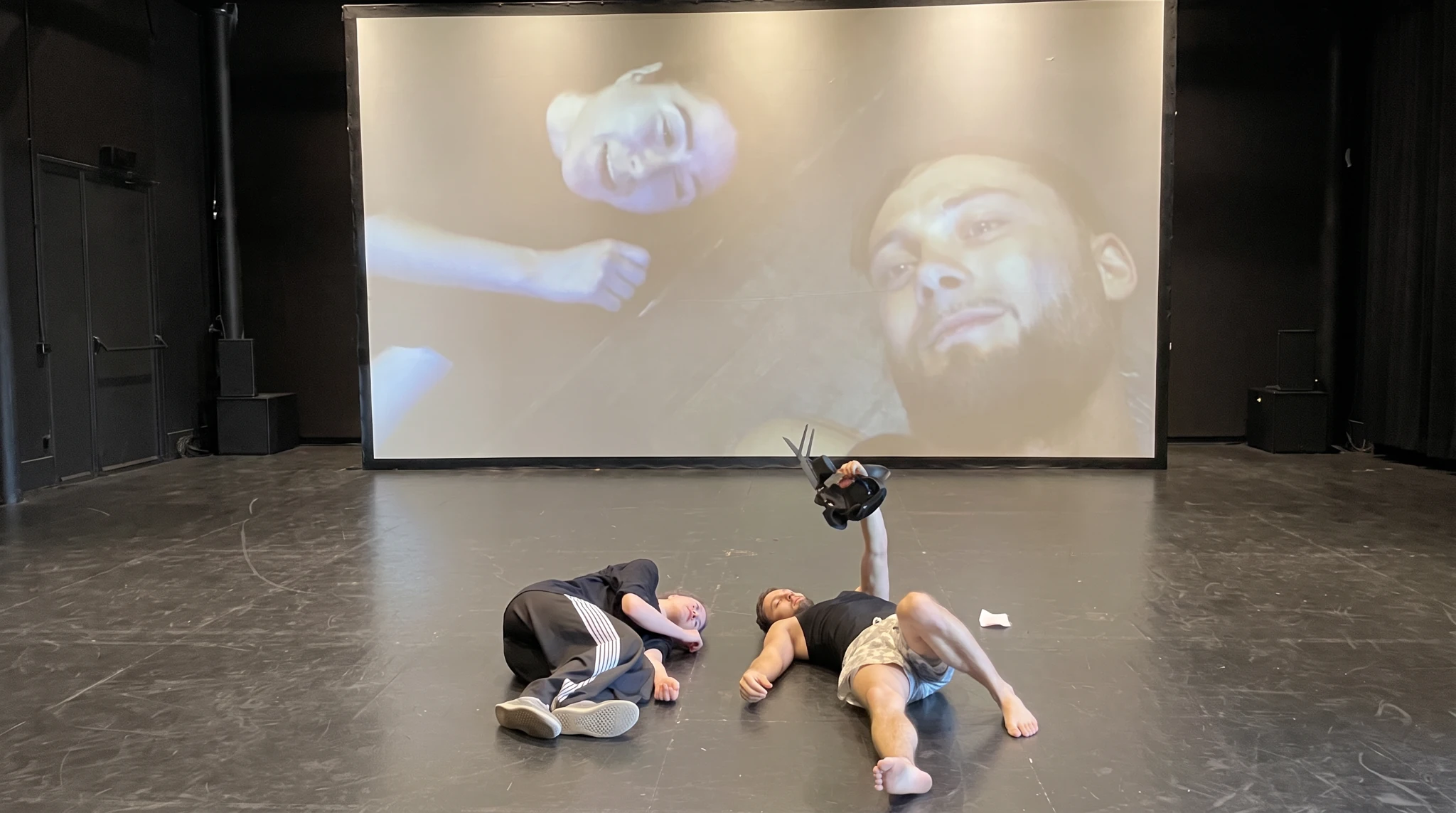 Deux interprètes allongés au plateau, une caméra à la main, avec leurs visages projetés sur un grand écran.